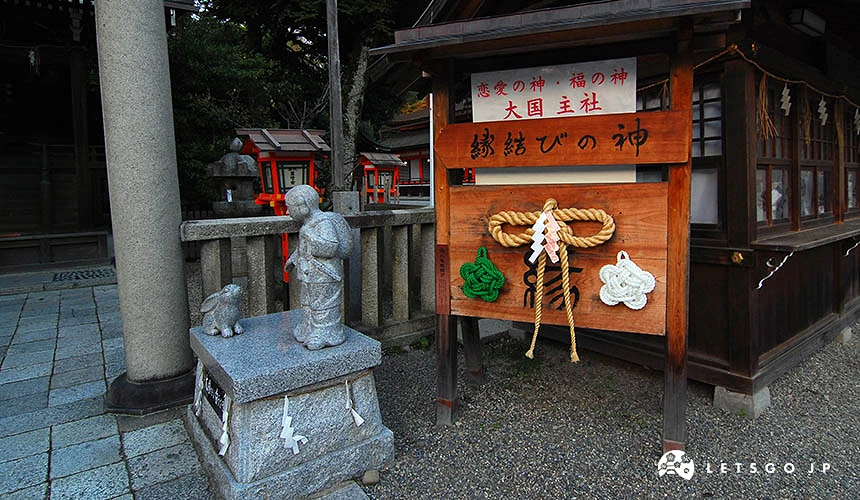 木製看板與石雕造像。木看板上寫有「恋愛の神・福の神 大国主社」與「縁結びの神」,並掛有注連繩與裝飾;前方左側為大國主命與因幡之白兔的石雕像。