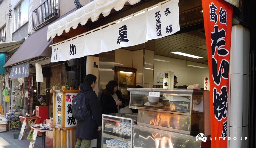人形町著名的百年鯛魚燒老店「柳屋」店面特寫。白色布幔上寫著黑色「柳屋」字樣,紅色的「高級鯛燒」旗幟十分顯眼。店門口有民眾正在排隊。