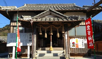 木造日式神社本殿入口,棕色瓦屋頂下懸掛大麻繩和鈴鐺,左右插有紅色旗幟,上書『寶當神社』字樣
