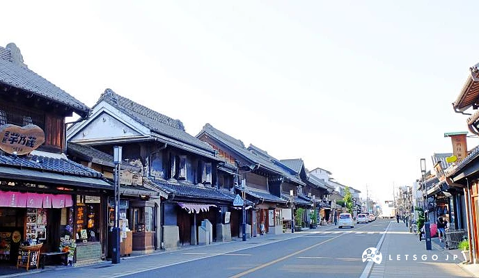 日本埼玉縣川越一番街藏造老街,街道兩旁排列著傳統黑瓦土藏造建築風格的店舖,包含掛有「芋乃藏」木製看板的商家與遠處行駛的車輛