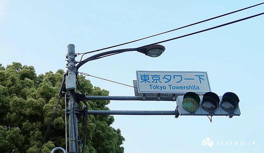 東京鐵塔下方的路口標示牌,顯示「東京タワー下(Tokyo Towershita)」字樣,旁邊是綠燈與街燈,背景為晴朗天空與茂密樹木