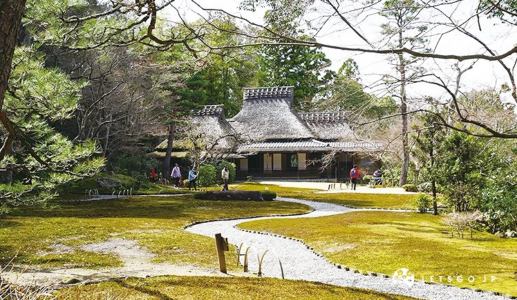 綠意盎然的吉城園庭園景觀,蜿蜒的碎石步道穿過長滿青苔的庭園,後方是一棟傳統的茅草屋頂日式建築
