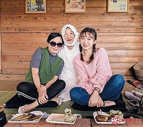 三名女性坐在木牆前,地面擺放食物,背景掛有框架