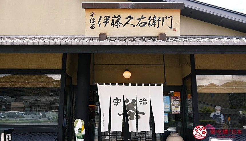 宇治老舖抹茶店正面入口,掛著白色暖簾,上方木製招牌書寫「伊藤久右衛門」字樣,門前擺放花卉裝飾。