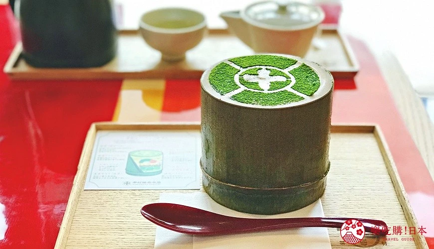 圓柱形竹製容器中盛裝抹茶甜點,表面撒滿細緻的抹茶粉並壓出環狀紋路;托盤上擺有紅色小木勺,背景可見茶壺與茶杯。