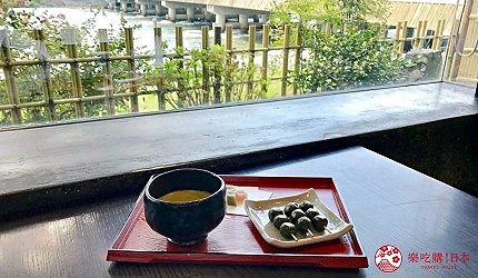 坐落於窗邊的木桌上,一只紅色托盤盛有一杯抹茶和一盤串狀團子,透過玻璃窗可見河畔與竹製柵欄景致。
