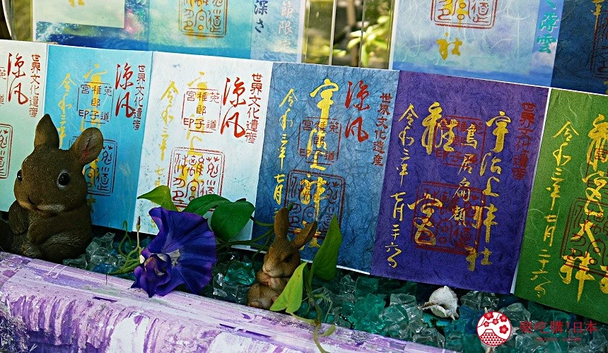 多張色彩繽紛的御朱印紙排列展示,上面蓋有紅色與金色朱印及手書書法,底下點綴藍紫色牽牛花與可愛兔子擺飾。
