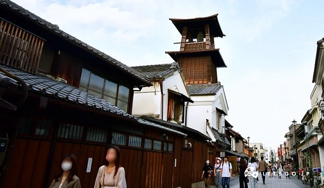 日本埼玉縣川越地標「時之鐘」木造鐘樓矗立於街道旁,兩側為傳統日式木造與藏造建築,街上有不少漫步觀光的遊客