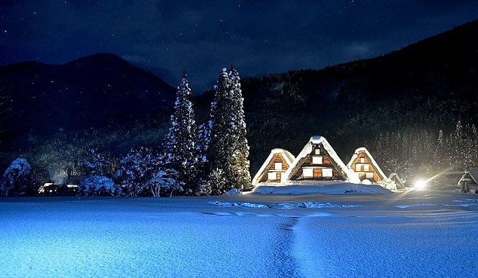 白川鄉合掌村的冬季點燈景色,雪地被藍光照亮,合掌造民屋在夜色中亮起暖光,呈現夢幻且富詩意的冬夜風景