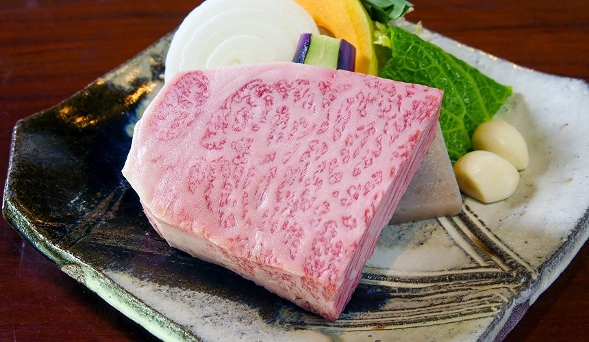 一塊油花濃郁的厚切佐賀牛生肉,旁邊擺有洋蔥切片、南瓜片、茄子、綠葉蔬菜和蒜瓣,盛放在日式陶瓷盤上。