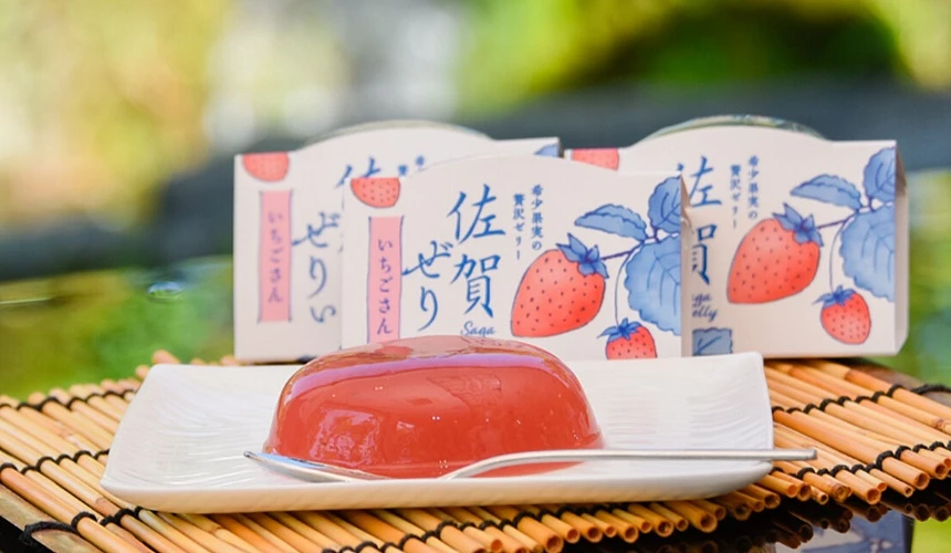 白色方盤上擺放著透亮鮮紅的草莓果凍,旁邊放著銀色小湯匙,後方則排列著印有草莓插畫與日文「佐賀ゼリー」字樣的包裝盒。