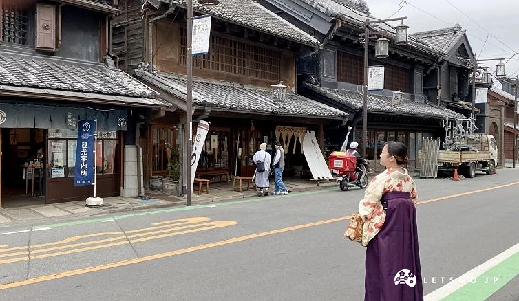 一位身穿和服與袴裝的女性站立於道路旁,背景為設有觀光案內所的傳統黑瓦土藏造建築與過往車輛