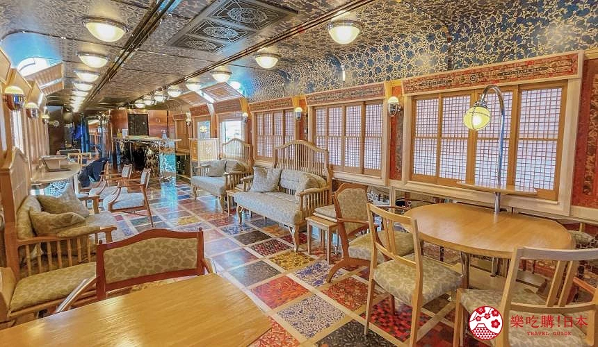 JR九州「雙星4047」觀光列車休憩車廂內部:彩色拼花地板與花紋天花板,兩側設有木質桌椅與沙發座椅,窗邊是日式木格窗,懸掛圓形燈具,整體營造典雅舒適的氛圍