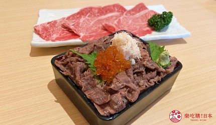 黑色方碗盛裝牛肉丼，配料有鮭魚卵、蟹肉絲與紫蘇葉，後方擺放生牛肉片