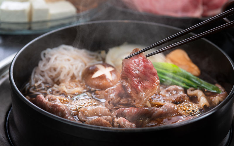 筷子夾牛肉片,壽喜燒鍋內有香菇、蔥與冒煙