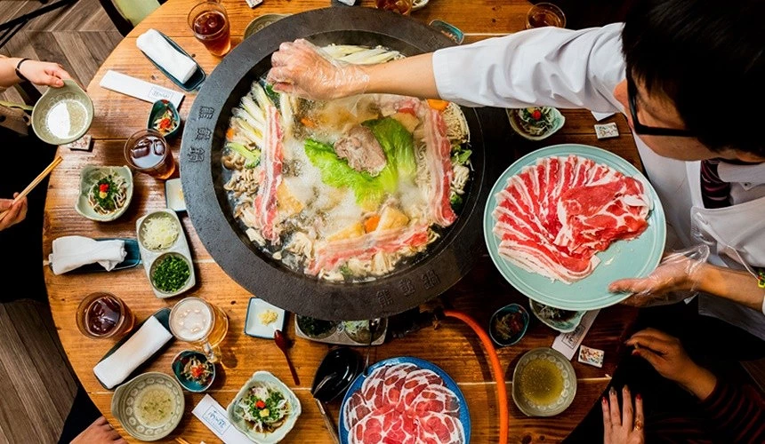 一個圓桌上有一鍋火鍋左邊有位男子拿著一排生肉要放入火鍋內桌上擺放著澎湃的小菜及肉片