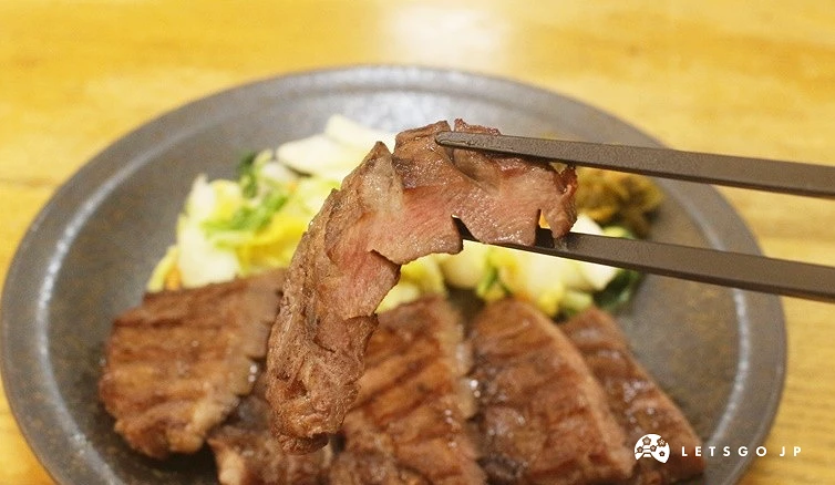深灰色圓盤前,一雙黑色筷子夾起一片厚切烤肉片。被夾起的肉片表面刻有深的交叉切痕,斷面呈現淡粉紅色的半熟熟度與柔軟肉質;背景的盤中擺放著其餘數塊微焦的厚切烤肉片,以及淺綠色與黃色的醃漬高麗菜配菜,下方為淡黃色的木質桌面。