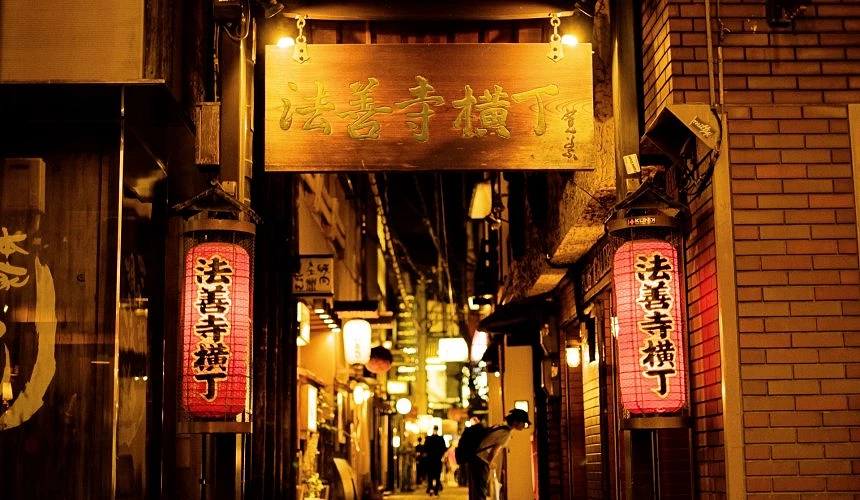 大阪法善寺橫丁夜晚街景,暖黃燈光映照下的傳統小巷,入口處掛有木製招牌與紅色燈籠