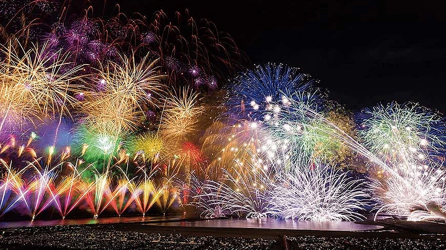 夜空中綻放的大型煙火秀,金色、紫色、藍色與粉色煙火同時施放,下方可見大量觀賞人群
