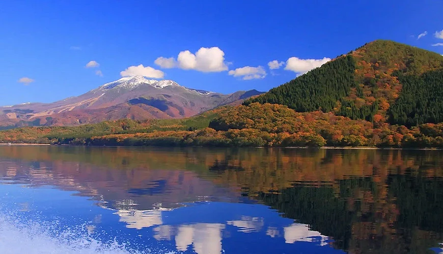 秋田縣田澤湖秋日風景,碧藍湖面倒映紅葉群山與遠方雪頂山峰