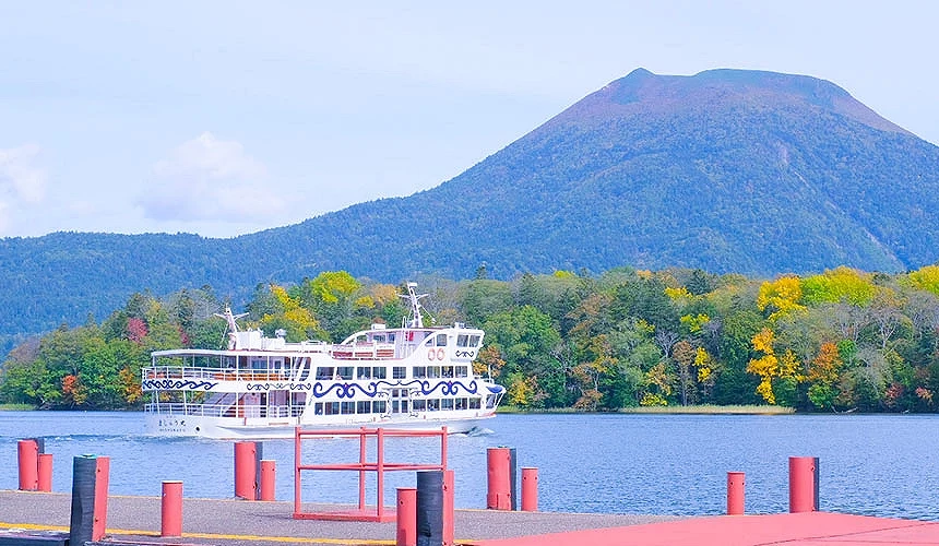 北海道阿寒湖秋季景色,遊覽船航行於湖面,背後群山與紅葉林相映成趣
