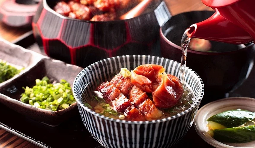 HITSUMABUSHI 花岡鰻魚茶泡飯,紅色茶壺正將高湯注入藍白條紋碗中,旁附青蔥、醃漬蔬菜與黑紅漆木桶碗