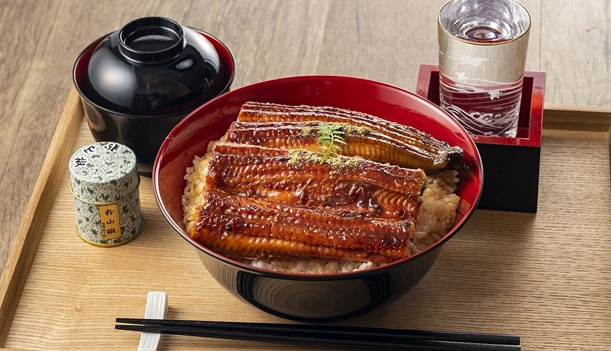 紅漆碗盛裝鰻魚飯,鰻魚片鋪滿米飯上方並淋有醬汁,旁附黑色湯碗、筷子與調味罐