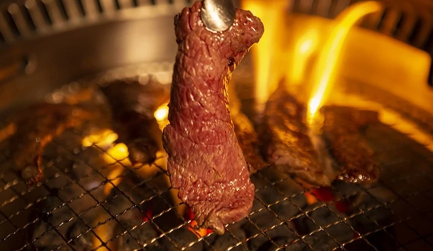 石用夾子夾起一片在炭火烤網上燒烤、油脂溢出的多汁燒肉。
