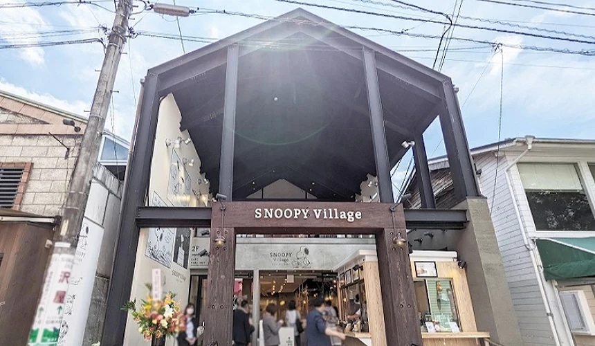 位於輕井澤的 SNOOPY Village 商店建築,擁有挑高的深色工業風鋼骨結構與巨大的斜屋頂設計,正門上方的深木色招牌印有 SNOOPY Village 字樣,店外裝飾有史努比插畫圖案與慶祝花籃