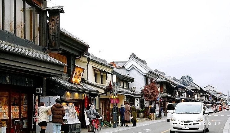 川越一番街藏造老街街景,道路兩旁為黑瓦與土藏造建築風格的傳統日式商鋪,街道上有行人與行駛中的白色汽車
