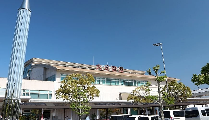 大分機場外觀,晴朗藍天下的白色建築外牆上可見「大分空港」紅色字樣,前方停有多輛車輛,畫面左側立有銀色造型裝置藝術,周邊有綠樹