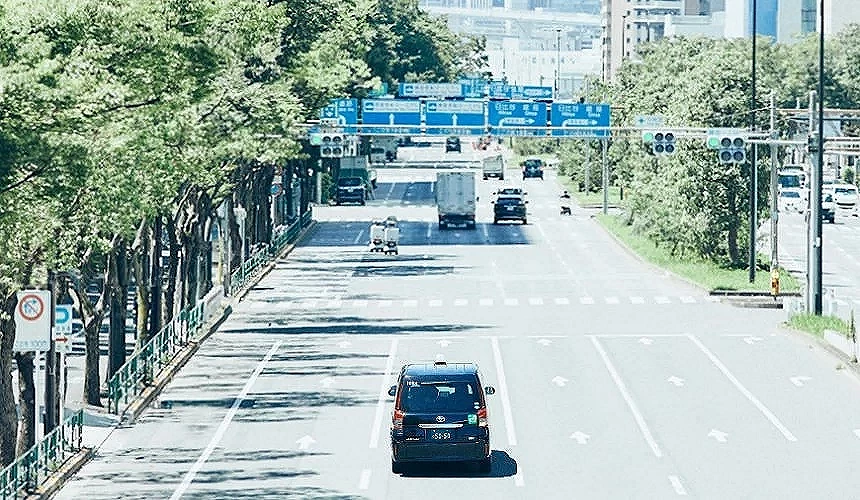 一輛日本計程車行駛在綠蔭大道上,前方為城市幹道與藍色交通標誌,展現順暢通勤的城市風景