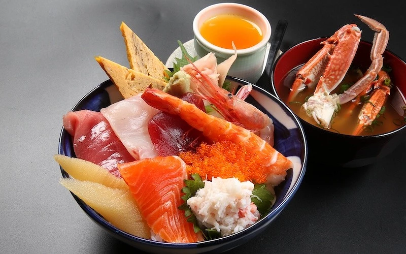 擺在桌面上的一碗裝有生鮭魚、魚卵、蟹肉絲等配料的丼飯，旁邊有布丁和盛滿蟹腳的湯
