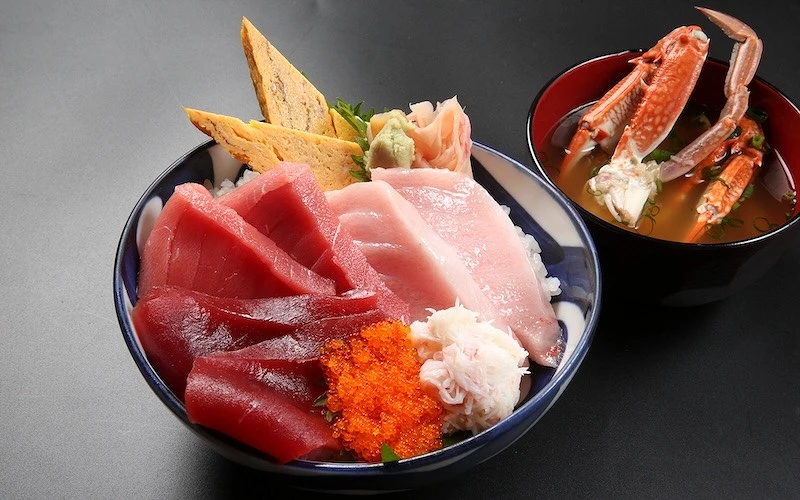 擺在桌面上的一碗裝有生鮪魚大腹、鮪魚中腹、鮪魚赤身等配料的丼飯，旁邊有盛滿蟹腳的湯