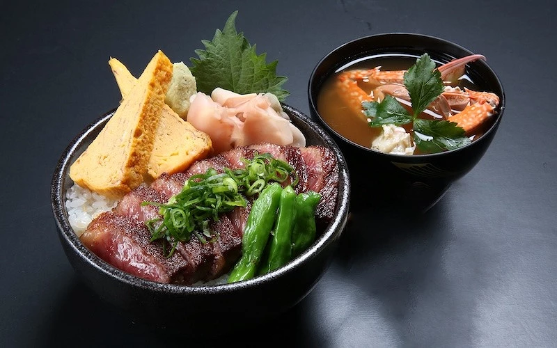 擺在桌面上的一碗裝有烤牛排、糯米椒、漬物、玉子燒的丼飯，旁邊有盛滿蟹腳的湯