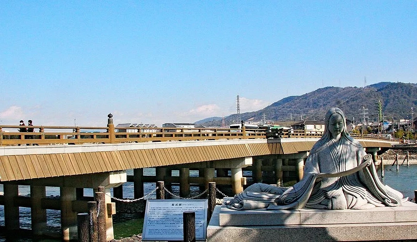 宇治川旁的紫式部雕像與橫跨河川的宇治橋,背景可見山巒與住宅,展現寧靜的古都風情