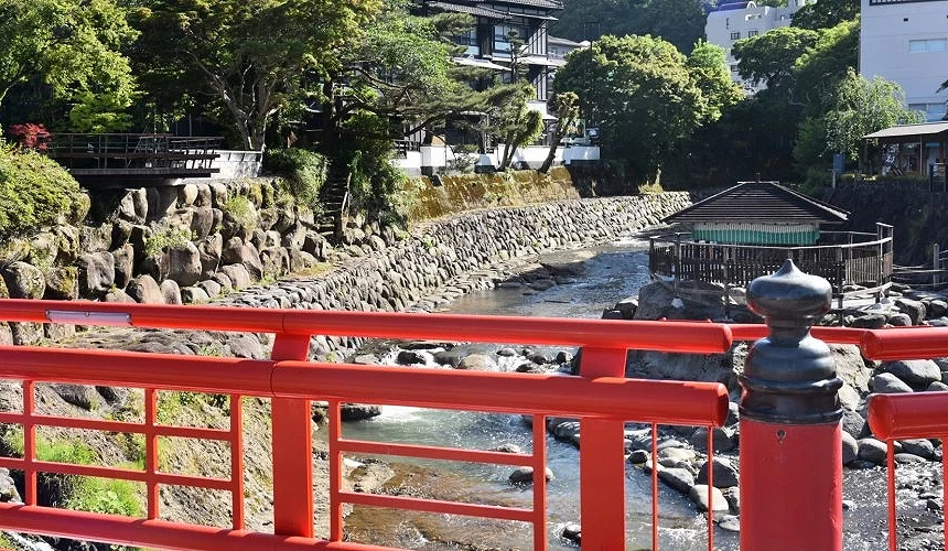 伊豆修善寺溫泉街的紅色欄杆橋與清澈溪流,背景是傳統日式建築與綠意盎然的自然景觀。