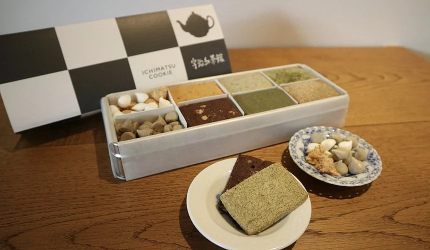 宇治紅茶館的招牌商品・イチマツクッキー(ICHIMATSU COOKIE),以各式抹茶、焙茶風味方形餅乾整齊排放於盒中,旁邊搭配茶點與精緻茶具。