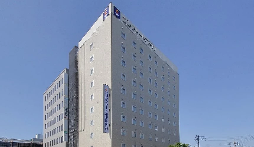 藍天映襯下的Comfort Hotel佐賀大樓,米色磚牆外觀上整齊排列多扇小窗,頂樓與立面淡藍色招牌標示飯店名稱
