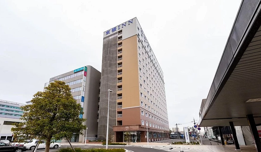 灰白天空下的東橫INN飯店佐賀站前,米色與咖啡色外牆交錯,頂樓標有藍字飯店招牌