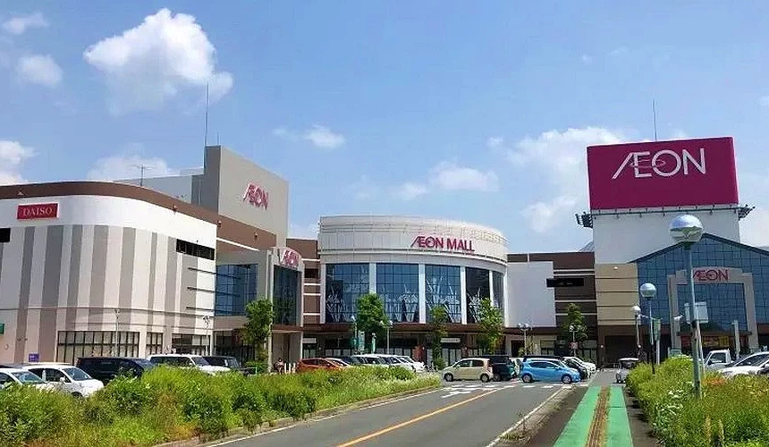 藍天白雲下,一條道路兩旁停滿汽車,盡頭為大型AEON購物中心,中央圓弧玻璃立面,右側高掛AEON紅色招牌,左側可見DAISO店招