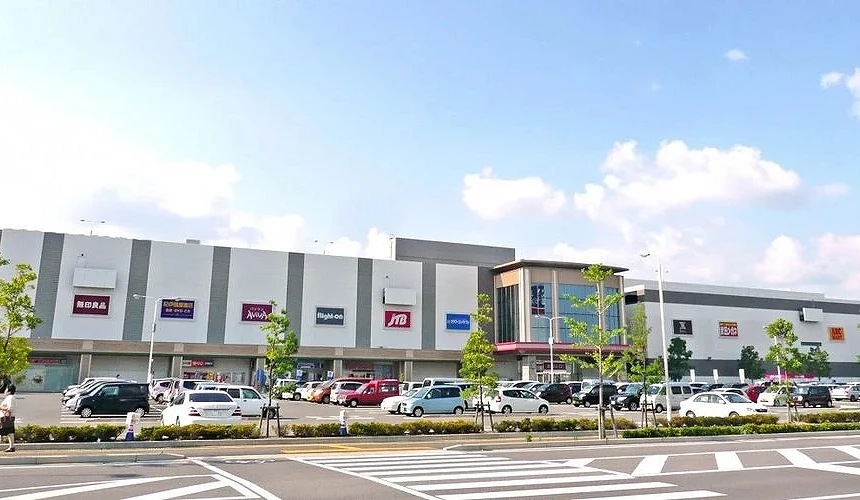 藍天白雲下的購物中心外觀,立面上有無印良品、JTB、Right-on等店鋪招牌,前方停車場停滿汽車,路旁有人行道