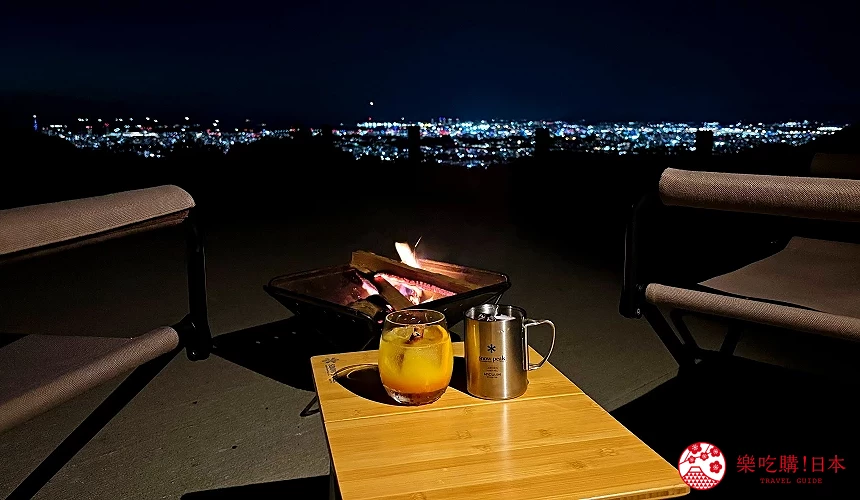 夜晚戶外座位前有營火,桌上放置果汁與不鏽鋼杯,遠處是城市夜景