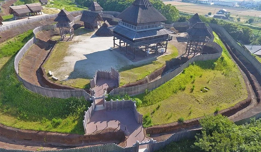 空中俯瞰吉野里遺跡:中央茅草屋頂的復原建築及多座瞭望台,四周以木製柵欄環繞並挖有護城溝渠,背景綠意田野延伸至遠方