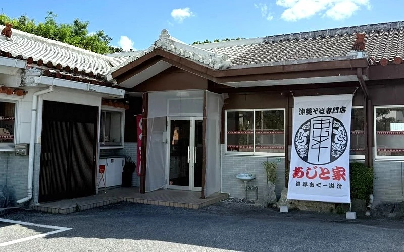 傳統屋瓦建築餐廳外觀,門口掛有「沖繩そば専門店 あじと家」布旗