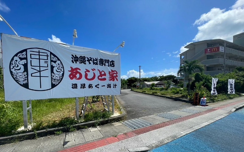 藍天下道路旁立牌,上寫「沖繩そば専門店 あじと家」