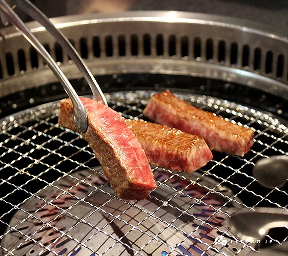 金屬夾夾起一塊正在燒烤的牛肉,烤網上還有兩塊肉