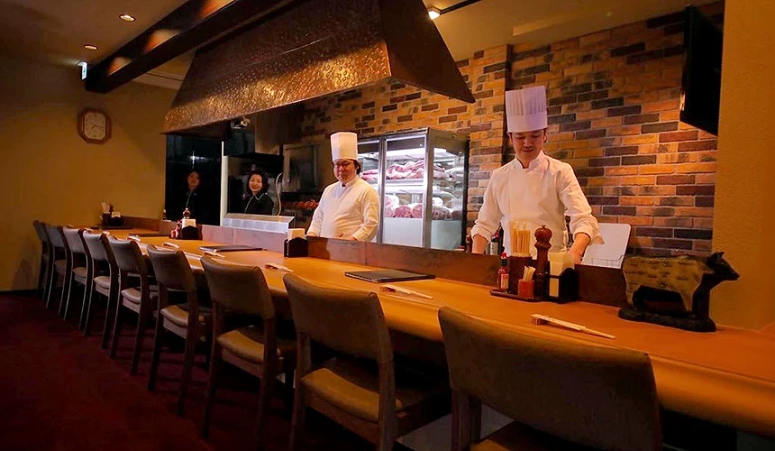 餐廳內吧檯座位前,兩位廚師站立,後方可見冷藏櫃中的牛肉