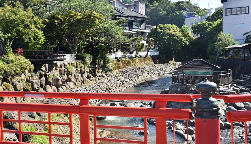 修善寺溫泉街的紅色欄杆橋與河畔「獨鈷之湯」溫泉設施,背景為傳統旅館與綠意盎然山林景觀