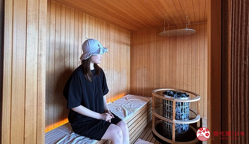 一名女子坐在木質蒸氣室長椅上,旁邊有裝滿石頭的加熱爐